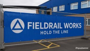 Wholesale mesh fence banners Fieldrail Works Hold The Line print on rectangular mesh panel zip-tied to industrial fence, iPopupbanner watermark